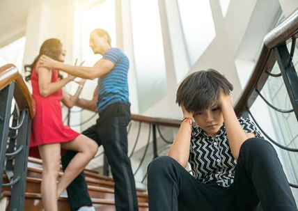 a child sitting on the stairs with his parents fighting behind him.